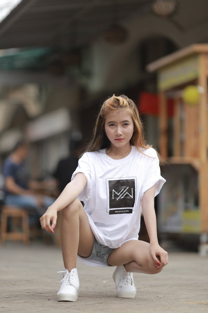 Girl in white t-shirt squatting outdoors, streetwear fashion pose.