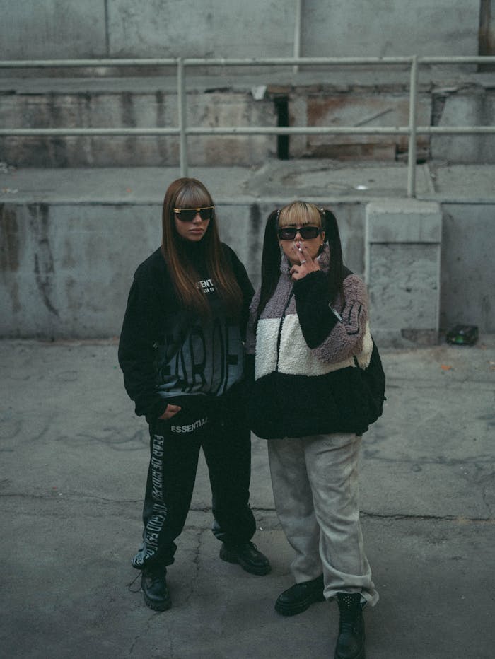 Two stylish individuals pose in an urban concrete setting with modern fashion.