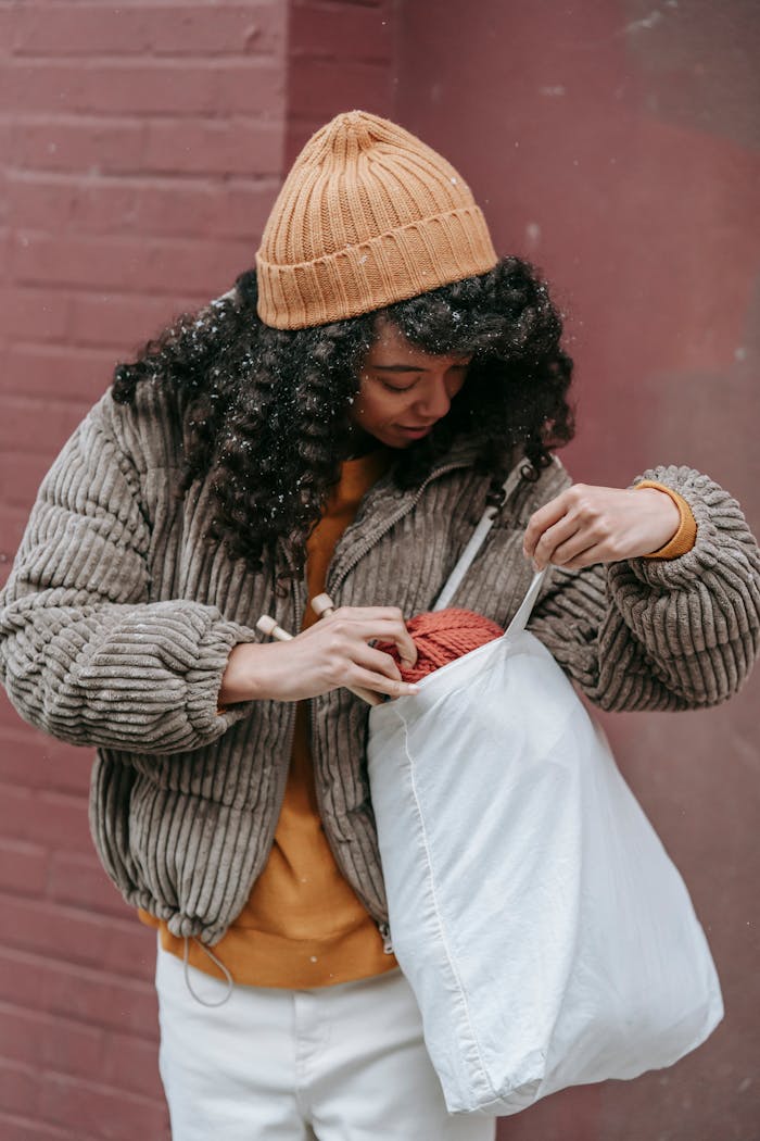 Home Young woman in winter attire examines yarn in eco-friendly bag against a brick wall.